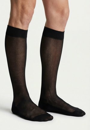 Männliche Beine, die durchsichtige schwarze knielange Socken mit solidem schwarzem Zehen- und Bündchenbereich auf hellem, schlichtem Hintergrund tragen.