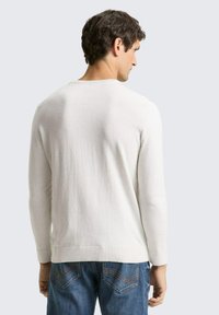 Hellbeiger Pullover aus weichem Stoff mit rundem Ausschnitt und langen Ärmeln, kombiniert mit blauen Jeans, die dekorative Nähte auf den Gesäßtaschen aufweisen.