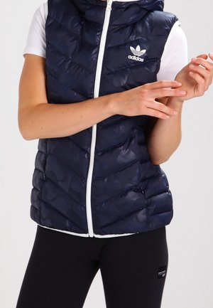Mouwloze navy Adidas donsvest met witte rits, gedragen over een wit t-shirt met korte mouwen, gecombineerd met zwarte legging.