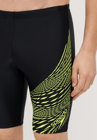 Μαύρο σορτς για κολύμπι με γεωμετρικό μοτίβο κίτρινου χρώματος στη αριστερή πλευρά. Φτιαγμένο από ελαστικό υλικό με μεσαίο μήκος και εφαρμοστή γραμμή.