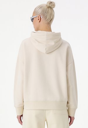 Personne aux cheveux blonds coiffés en chignon portant un sweat à capuche beige clair et un pantalon assorti, de dos, portant des lunettes de soleil grises sur fond blanc.