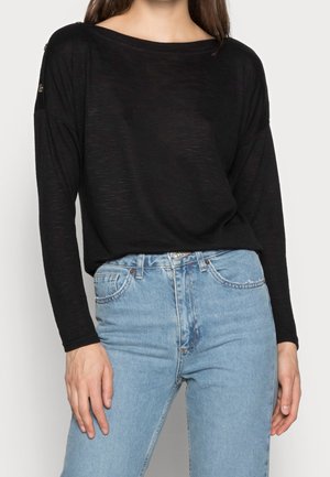 Black long-sleeve top with a relaxed fit, featuring a wide neckline and decorative buttons on the shoulders, paired with light blue high-waisted jeans.