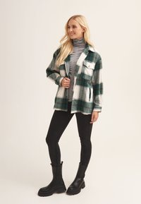 Green and white plaid flannel jacket with two front pockets, layered over a gray turtleneck, paired with black leggings and knee-high boots.