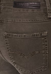 Jeans de mezclilla verde oliva con un bolsillo trasero con costura decorativa y un logotipo de cuero de la marca encima de la cinturilla. Tela texturizada.