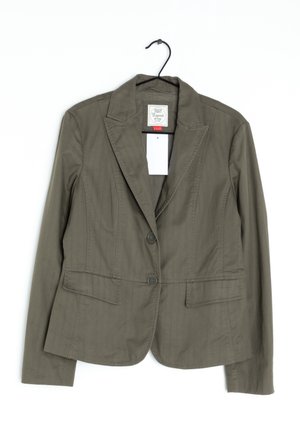 Blazer da donna verde oliva con colletto a risvolto, due bottoni, tasche anteriori a patta, appeso a un gancio nero su sfondo bianco.