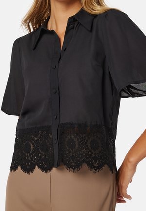 Blouse noire à manches courtes avec boutons et bordure en dentelle florale à l'ourlet, portée avec un pantalon beige, montrant le haut du torse et le bras droit reposant sur le côté.