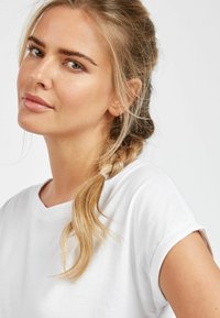 Valge ümmarguse kaelusega T-särk, mille varrukad on rullitud, kombineeritud lahtise palmikuga. Sile tekstuur, vabaaja disain ja neutraalne värv rõhutavad lihtsust.