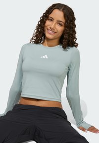 Jasnoniebieska sportowa koszulka z długim rękawem, okrągłym dekoltem i białym logo na klatce piersiowej. Tkanina jest gładka, a fason dopasowany.