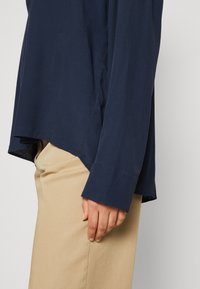 Camicia a maniche lunghe blu navy in tessuto morbido, con orlo curvo e polsini aderenti, abbinata a pantaloni beige, con la mano appoggiata delicatamente sul fianco.