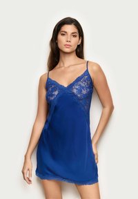 Chemise en soie bleue avec des accents en dentelle, décolleté en V profond et fines bretelles réglables. Sa texture douce et sa légèreté améliorent le confort.