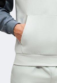 Άτομο που φοράει δίχρωμο φούτερ με κουκούλα, με το ένα χέρι στην μπροστινή τσέπη, δείχνοντας την υφή και τις ραφές του ανοιχτού και σκουρού υφάσματος.
