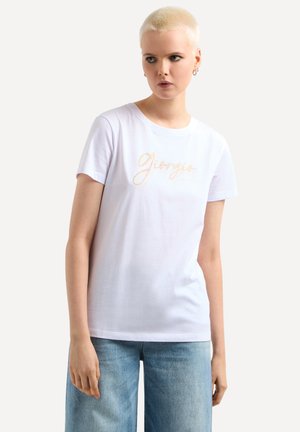 T-shirt bianca in cotone con maniche corte, caratterizzata dalla scritta "Giorgio" in un font script luccicante. Abbinata a jeans a gamba larga di colore blu chiaro.