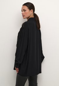 Blusa nera oversize in tessuto morbido, con dettagli in pizzo intricati sulle spalle e una vestibilità rilassata con polsini con bottoni.