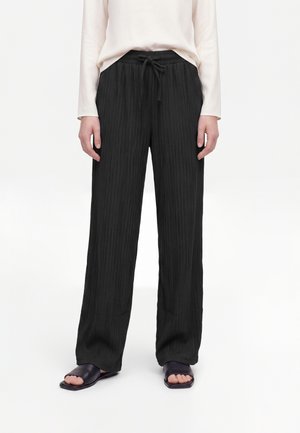 Persoon draagt zwarte, wijde geplooide broek met een aantrekkoord in de taille, een licht shirt met lange mouwen en zwarte sandalen met open tenen.