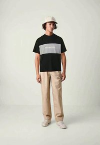 Young man wearing a black and gray Jack & Jones t-shirt, beige pants, white sneakers, and a white cap, standing against a plain background.