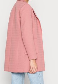 Giacca rosa a motivo spina di pesce con una vestibilità ampia, colletto a scendere e due tasche frontali. Indossata sopra un denim azzurro chiaro.