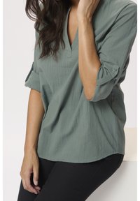 Blusa verde chiaro, texture, con scollatura a V, maniche arrotolate con bottoni, indossata con pantaloni neri. Design semplice e dalla vestibilità rilassata.