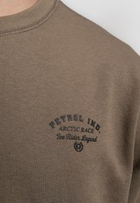 Bruine katoenen sweatshirt met een ronde hals, geribbelde afwerking en een contrasterend zwart logo met tekst op de linkerkant van de borst.