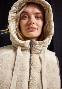 Gilet couleur crème avec capuche, doté d'une texture en sherpa duveteux et de panneaux en tissu lisse. Fermeture éclair avec tirette de marque et bretelles étroites.