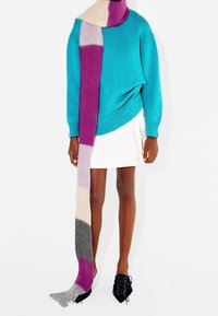 Turkusowy sweter o prążkowanej fakturze, w zestawieniu z długim, wielokolorowym szalikiem w odcieniach różu, fioletu, szarości i kremu; biała spódnica i czarne buty.