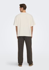 Jasnobeżowa, luźna koszulka z krótkim rękawem i okrągłym dekoltem; w połączeniu z ciemnobrązowymi spodniami o szerokich nogawkach oraz beżowymi butami. Widok od tyłu.