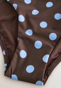 Tissu marron avec de grands pois bleu clair espacés de manière régulière, présentant des coutures froncées sur les bords, probablement une partie d'un maillot de bain ou de vêtements.