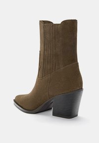 Bottine en suède marron avec un bout effilé, des accents nervurés et un talon empilé. Le design présente une texture lisse et un minimalisme dans les détails.
