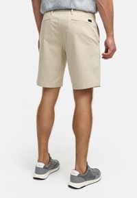 Beige bomuldsshorts med en afslappet pasform, udstyret med baglommer og en glat tekstur, parret med grå sneakers med hvid accent.