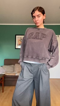 Szürke, rövidített pulóver "GAP" logóval és túlméretezett ujjakkal, széles szárú szürke nadrággal párosítva. Egyszínű háttér, fa padló.