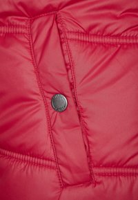 Interior de chaqueta roja acolchada con textura tipo acolchado, tejido suave, un prominente botón de presión negro redondo y dos bolsillos visibles.