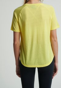 Osoba ubrana w luźną, krótką, żółtą siateczkową bluzkę z krótkim rękawem i dopasowane czarne spodnie, widziana od tyłu na jednolitym tle.