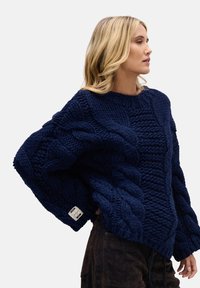 Granatowy sweter dziany z dużymi wzorami warkoczowymi, szerokimi rękawami i okrągłym dekoltem. Materiał wydaje się gruby i teksturowany.