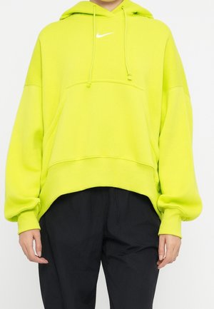 Person iført en lys gul-grøn Nike hættetrøje med snore og sorte bukser, vist fra nakke til hofter mod en ensfarvet baggrund.