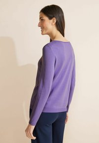 Lila stickad tröja med långa ärmar och rundad halsringning. Slät textur, ribbat fåll och en avslappnad passform. Bärs över mörka byxor.