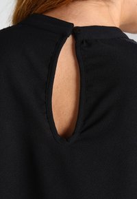 Blouse noire avec une ouverture en forme de clé à l'arrière, encolure ronde et tissu texturé. Elle dispose d'une fermeture à bouton à la nuque.