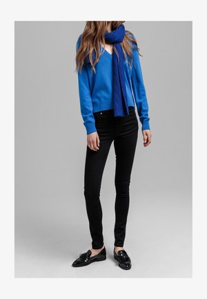 Blauwe gebreide trui met V-hals, gecombineerd met zwarte slim-fit jeans. Een bijpassende blauwe kabelgebreide sjaal en glanzende zwarte loafers maken de look compleet.