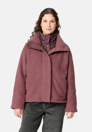 Frau mit lockigem Haar, die eine mauvefarbene Fleecejacke über einer wattierten Jacke und dunklen Hosen trägt, steht vor einem einfarbigen hellen Hintergrund.