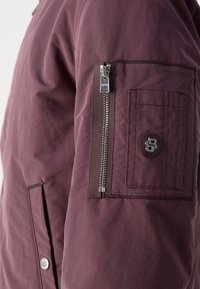 Maroon jacket featuring a zippered sleeve pocket, button accents, and a smooth texture. Includes a logo on the pocket and stitching details.