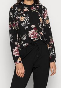 Schwarze Bluse mit floralem Muster in Rot, Orange und Weiß, mit langen Ärmeln und gerafftem Ausschnitt. Kombiniert mit schwarzen Hosen.