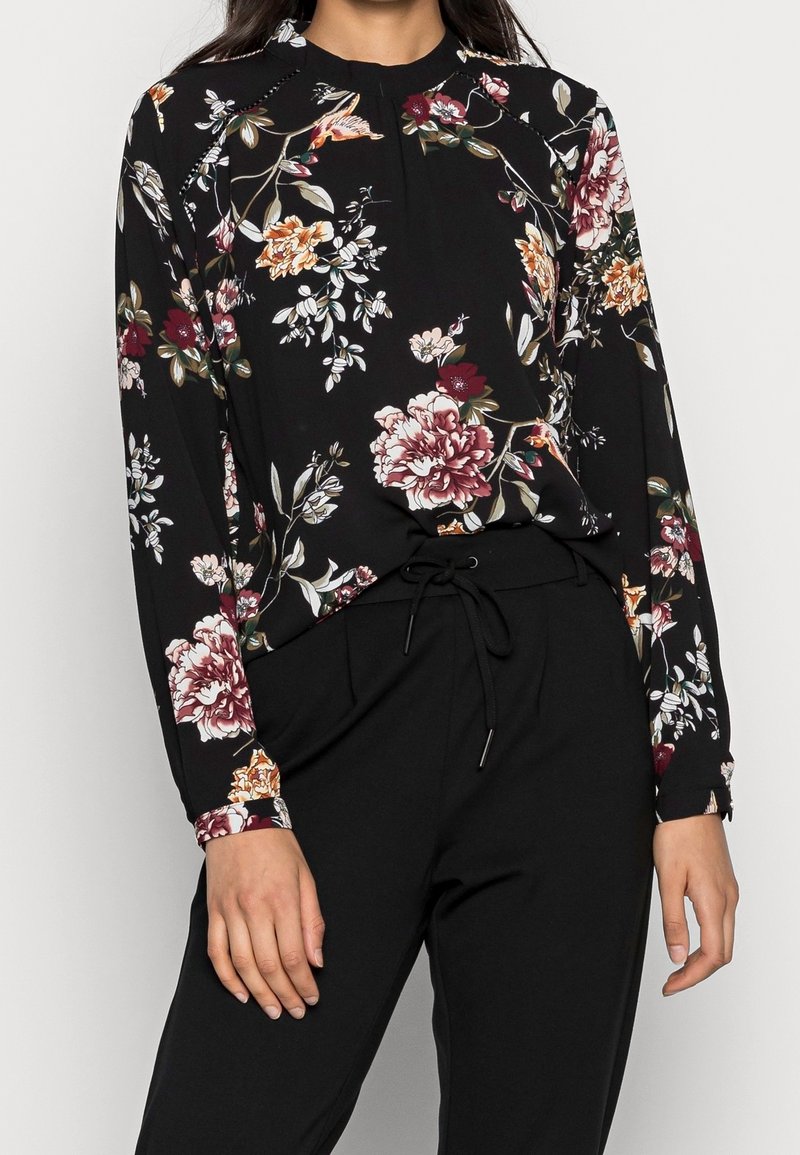 Schwarze Bluse mit floralem Muster in Rot, Orange und Weiß, mit langen Ärmeln und gerafftem Ausschnitt. Kombiniert mit schwarzen Hosen.
