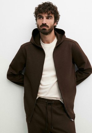 Uomo con capelli ricci e barba che indossa una felpa marrone scuro con cerniera e pantaloni abbinati, in piedi con le mani dietro la schiena contro un muro bianco.