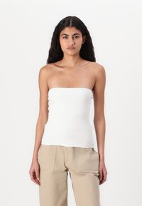 Hvit stroppeløs topp laget av glatt stoff, tilpasset design, med rett halslinje. Bært med lysebeige bukser, minimale detaljer.