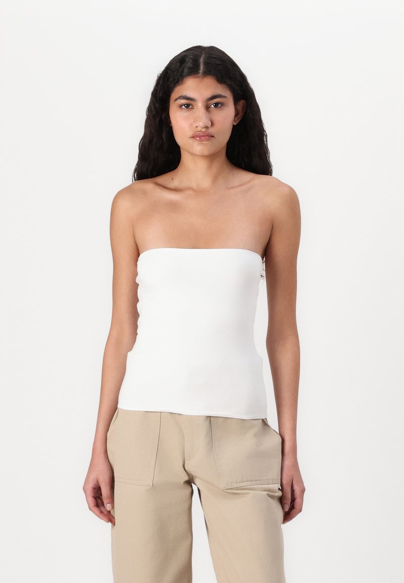 Hvit stroppeløs topp laget av glatt stoff, tilpasset design, med rett halslinje. Bært med lysebeige bukser, minimale detaljer.