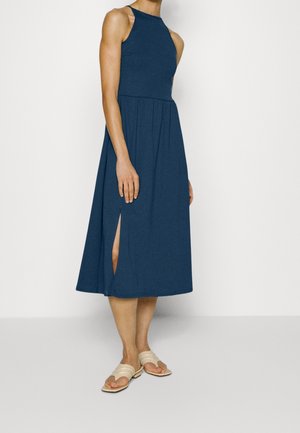 Bez rękawów, granatowa sukienka midi z miękkiej tkaniny, z wysokim dekoltem, rozkloszowanym dołem i bocznym rozcięciem. Dopełniona beżowymi sandałami na płaskiej podeszwie.