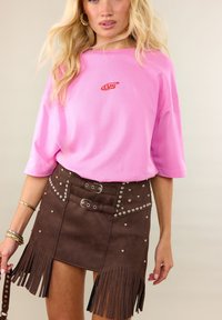 Femme portant un t-shirt rose ample avec un logo rouge et une mini-jupe marron cloutée avec double ceinture et détails à franges.
