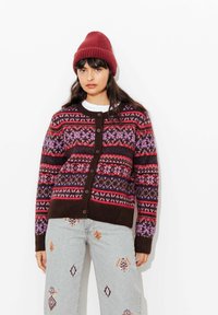 Cardigan en maille marron foncé avec des rayures à motifs multicolores, doté de boutons sur le devant. Associé à un jean clair et un bonnet rouge.
