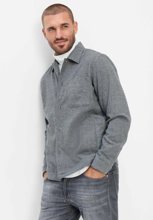 Mand med kort hår og skæg iført grå knappet jakke og grå jeans, stående med hænderne i lommerne og ser til siden.