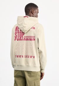 Sudadera beige con un gran estampado gráfico en la parte posterior en rosa oscuro, con el texto "NUNCA DEJES DE EXPLORAR" y las coordenadas debajo. Cuenta con una capucha con cordones.