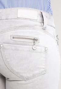 Jean en denim gris clair avec une poche zippée, des coutures visibles et une étiquette décorative de marque sur la taille. Texture lisse.