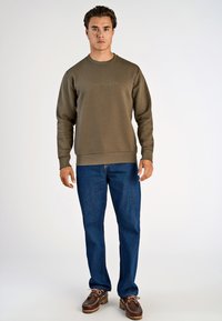 Felpa verde oliva con collo rotondo, polsini e orlo a costine. Abbinata a jeans blu e scarpe marroni con lacci e suole testurizzate.